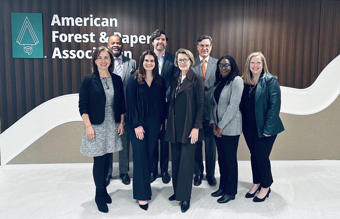 8 people standing in a cluster smiling in front of a sign that reads "The American Forest & Paper Association"