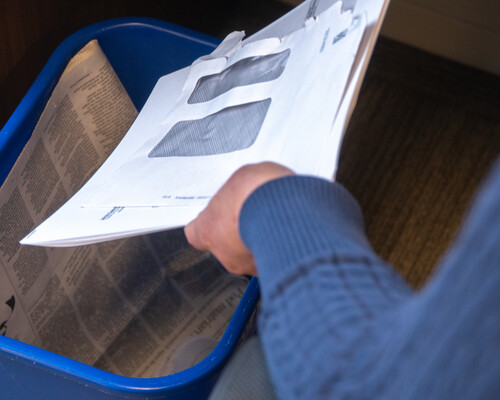 A person putting mail and a newspaper in a recycling bin.